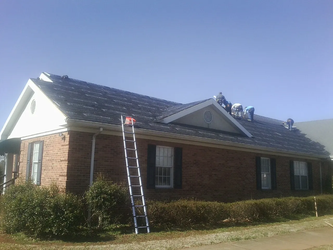 Crew tearing shingles off a brick ranch roof with a ladder set — Metal Roof Repair work by our crew in Toano