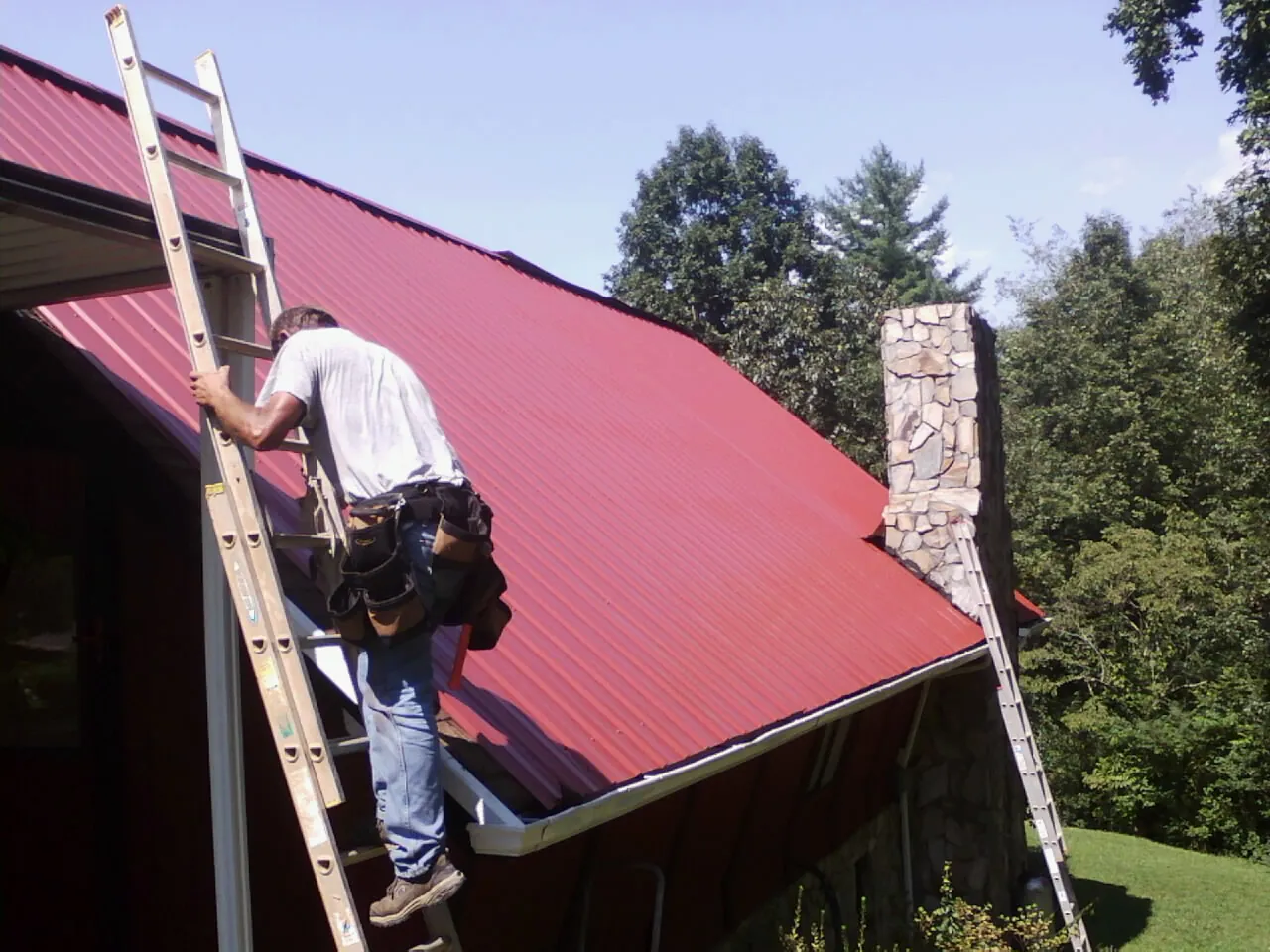 Roofer on a ladder beside a red metal roof and stone chimney — Soffit & Fascia Repair by Roofing Brothers in Toano
